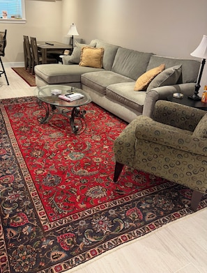 Ein Wohnzimmer mit Ecksofa, einem gemusterten Sessel, einem Glastisch und einem roten und schwarzen Teppich.