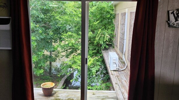 Een kamer met een schuifdeur naar een balkon met uitzicht op groen. Er staat een houten tafel en stoelen in de kamer.