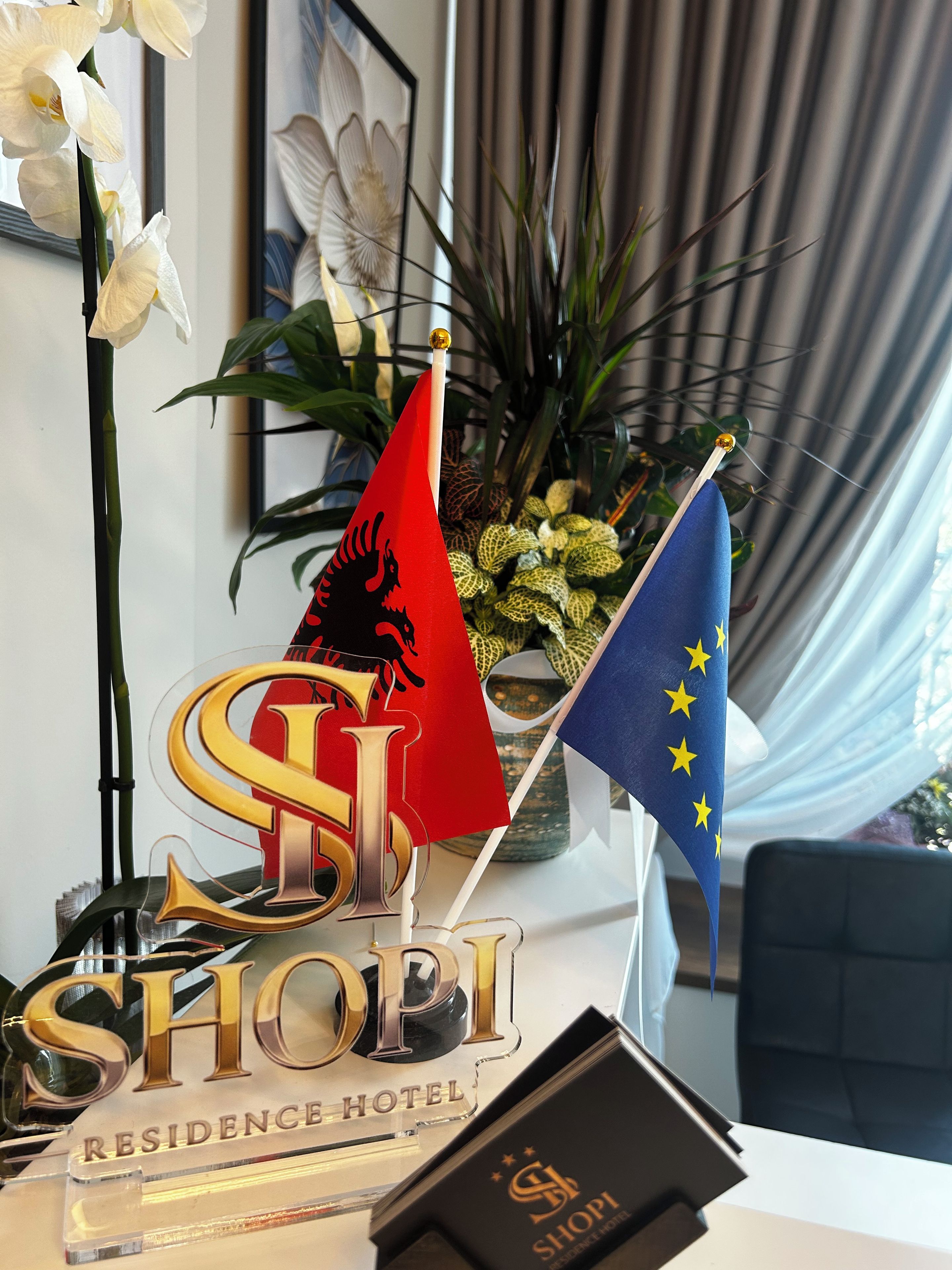 Un mostrador de recepción con banderas, un letrero que dice 'SHOPI RESIDENCE HOTEL' y una planta en maceta.