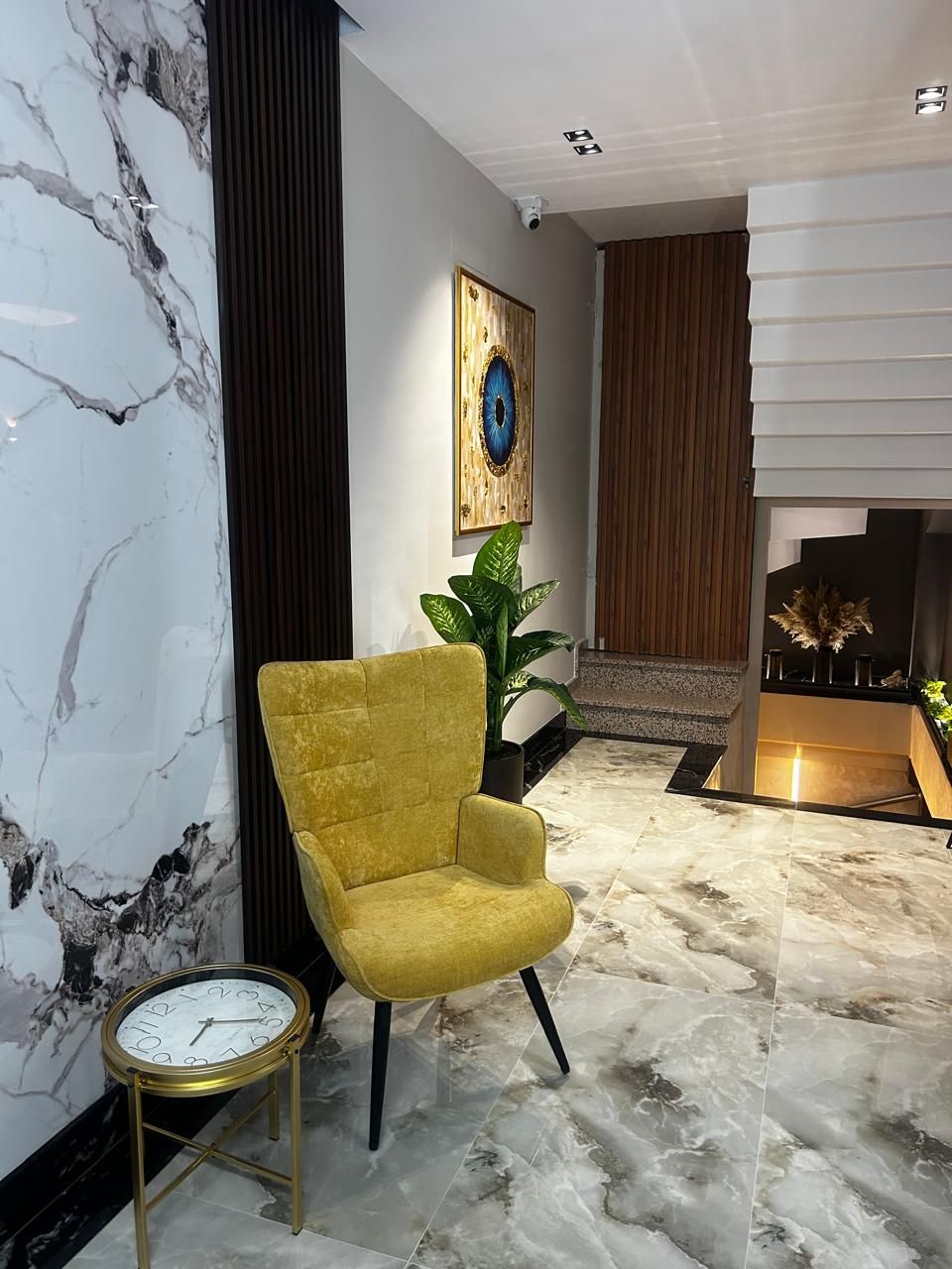 Un hall d’entrée moderne avec un fauteuil jaune, un mur en marbre et une horloge posée sur un piédestal.