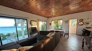 A spacious living area with a large sofa, a dining table, and a TV. There are large windows offering a view of the sea. - Top View Cottage in Lake Arenal (Río Chiquito)