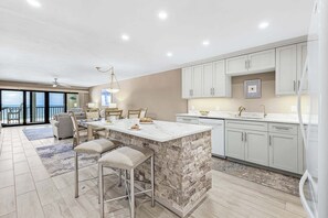 A modern kitchen with a central island, bar stools, a dining area with a view, and light-colored cabinetry. - Beachfront Sunsets, Heated Pool & Gulf Views Escape - Dolphin Watch 203 - Roelens (Fort Myers Beach)