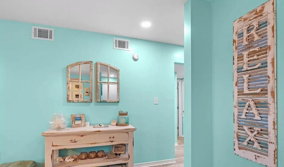 A room with teal walls, a vintage mirror, a decorative sign, and a wooden dresser with drawers.