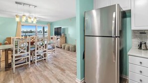 A modern kitchen with a stainless steel refrigerator, a dining area with wooden chairs, and a view through the window. - A14 Sundown Riverside Condominiums (Perdido Key)