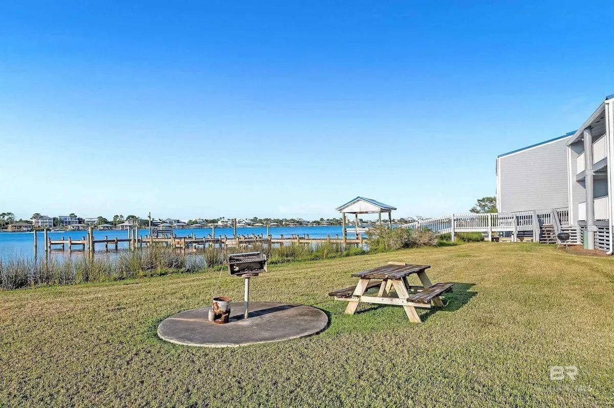 A grassy area with a barbecue grill, picnic table, and a gazebo overlooking a body of water.