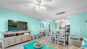 A living room with a TV, wooden entertainment unit, dining table with chairs, and a ceiling fan. - A14 Sundown Riverside Condominiums (Perdido Key)
