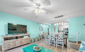 A living room with a TV, wooden entertainment unit, dining table with chairs, and a ceiling fan. - A14 Sundown Riverside Condominiums (Perdido Key)