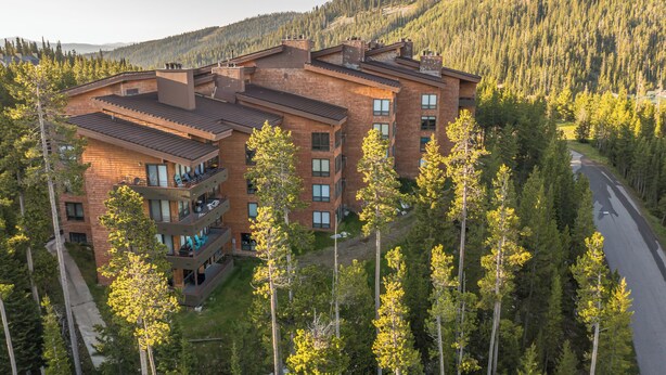 A multi-story hotel with a brick facade, multiple balconies, and surrounded by a forested mountain area. - NEW! Ski. Hike. Fish. Big Sky Mountain Village (Big Sky)