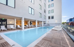 A modern hotel with a rooftop pool, lounge chairs, and a glass railing overlooking the city.