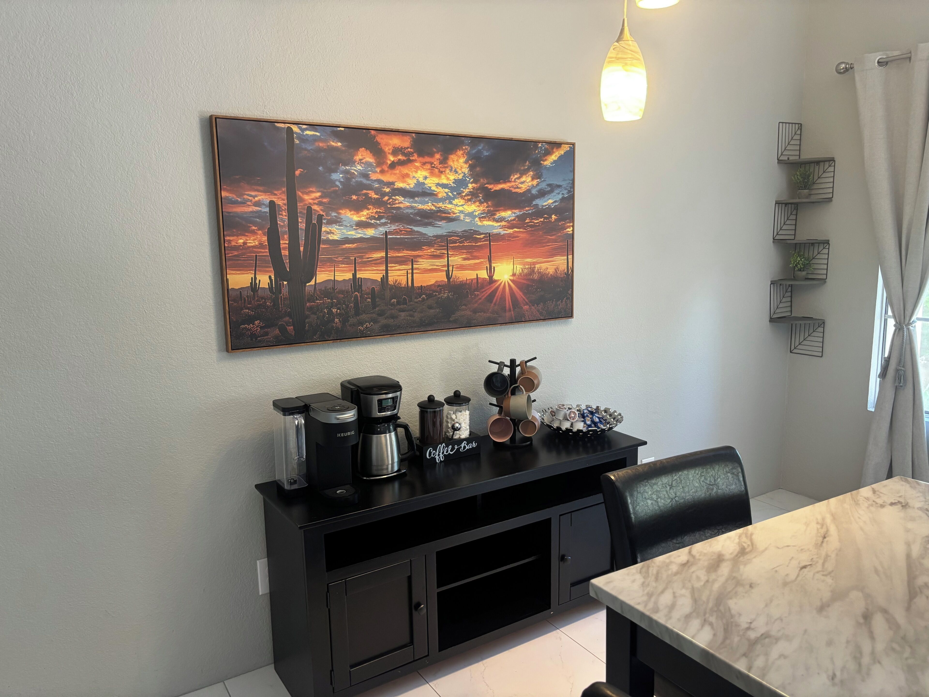 Un coin repas comprenant une table, des chaises, un espace dédié au café et un tableau représentant un coucher de soleil dans le désert, accroché au mur.