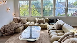 Un salon comprenant un canapé d’angle, une table basse en verre et une grande fenêtre donnant sur des arbres et des maisons.