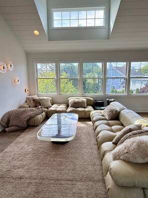 Ein Wohnzimmer mit einem Ecksofa, einem Glastisch und einem großen Fenster mit Blick auf Bäume und Häuser.