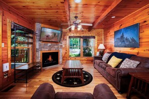 Ein gemütliches Blockhäuschen im Inneren mit Kamin, Holzwänden, Deckenventilator, Fernseher und großem Fenster mit Blick auf Bäume.