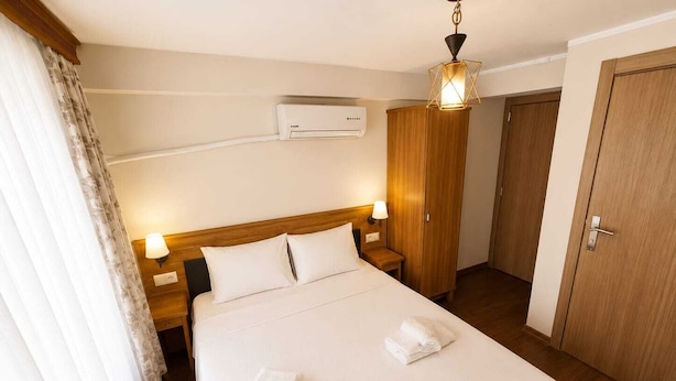 A hotel room with a bed, two bedside lamps, a wardrobe, an air conditioning unit, and a door. - Nisa Pansiyon (Ayvalık)