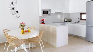 Een moderne keuken met witte kasten, een centraal eiland, een magnetron, een koffiezetapparaat, en rieten stoelen rond een witte tafel met een fruitschaal en bloemen.