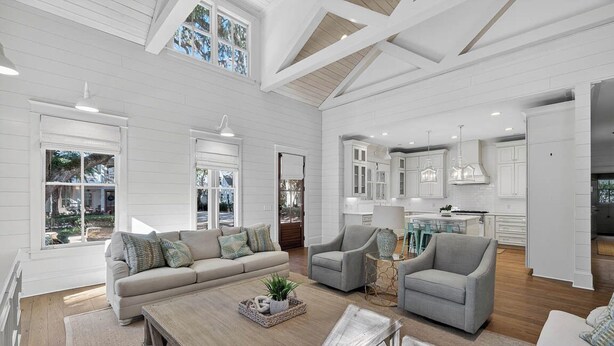 A spacious living room with a large sofa, a coffee table, and a fireplace. There are multiple windows and a kitchen area visible in the background. - Village Oaks Escape | Home steps to the Village! (St Simons Island)
