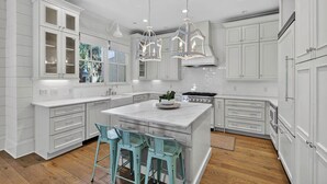 A modern kitchen with white cabinetry, a large island, pendant lights, and bar stools. - Village Oaks Escape | Home steps to the Village! (St Simons Island)