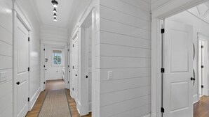 A hallway with white paneled walls, wooden flooring, and a door with a black handle. - Village Oaks Escape | Home steps to the Village! (St Simons Island)