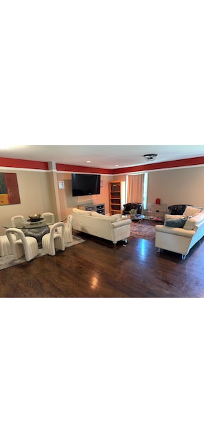 A spacious living room with a wooden floor, white sofas, a glass coffee table, and a flat-screen TV mounted on the wall. -     Relax & Unwind Stylish Decatur Home Near Downtown ATL (Decatur)