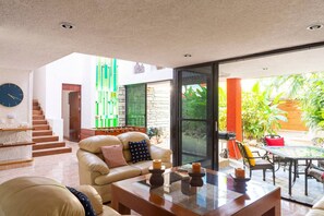 Ein geräumiger Wohnbereich mit einer Couch, einem Couchtisch und einem Kicker. Eine Treppe führt in ein Obergeschoss, und eine Glastür öffnet sich zu einer Terrasse mit Pflanzen.