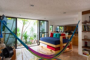 Ein Zimmer mit Hängematte, einem Bett mit bunten Kissen, einer Küchenzeile und großen Fenstern mit Blick auf Grünflächen.