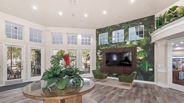 Um hall amplo com uma mesa de vidro redonda, cadeiras verdes, uma televisão grande e um mural tropical.