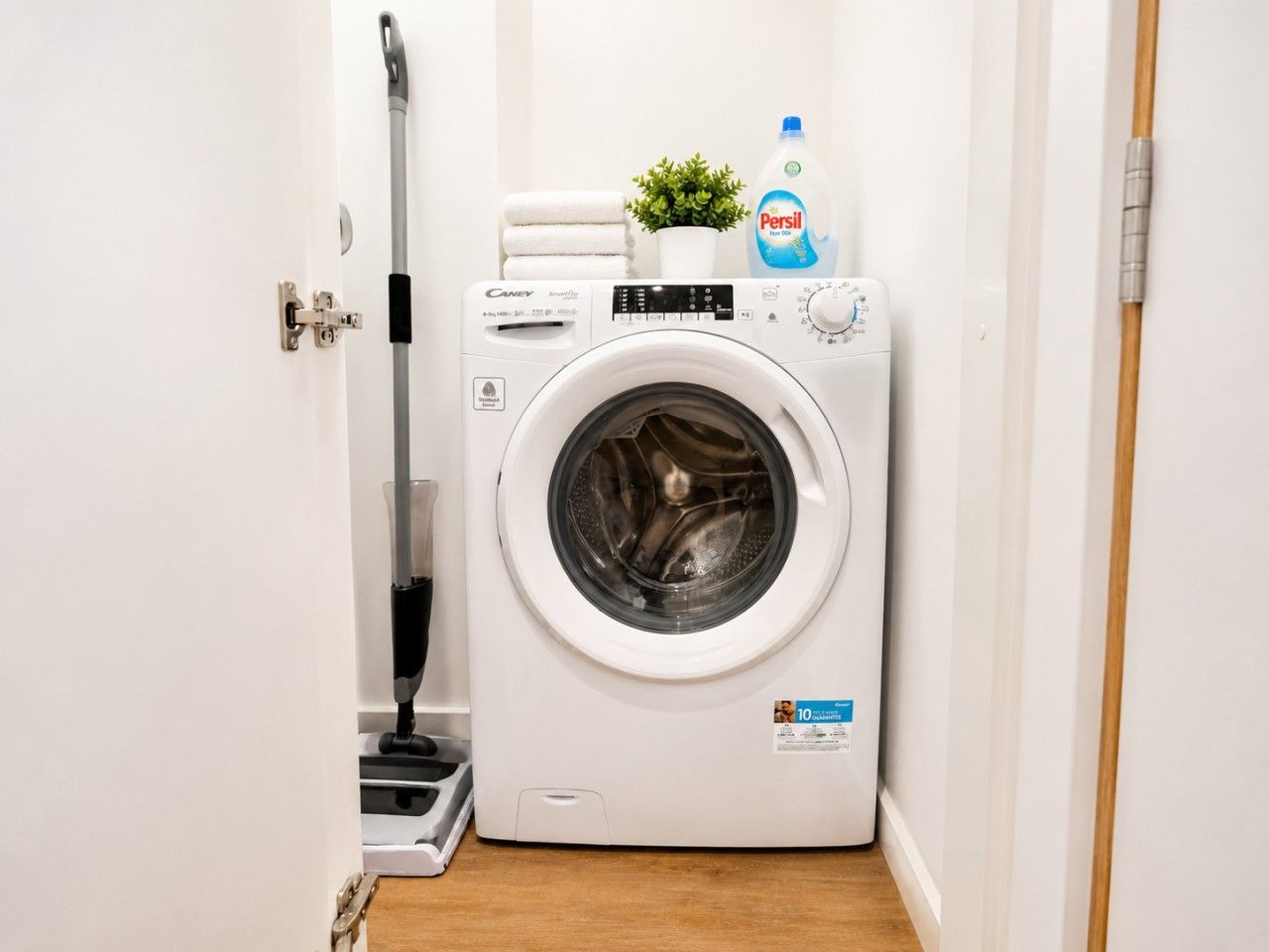Lavadora blanca compacta en un pasillo estrecho, con una pequeña planta en maceta y una botella de detergente Persil encima.