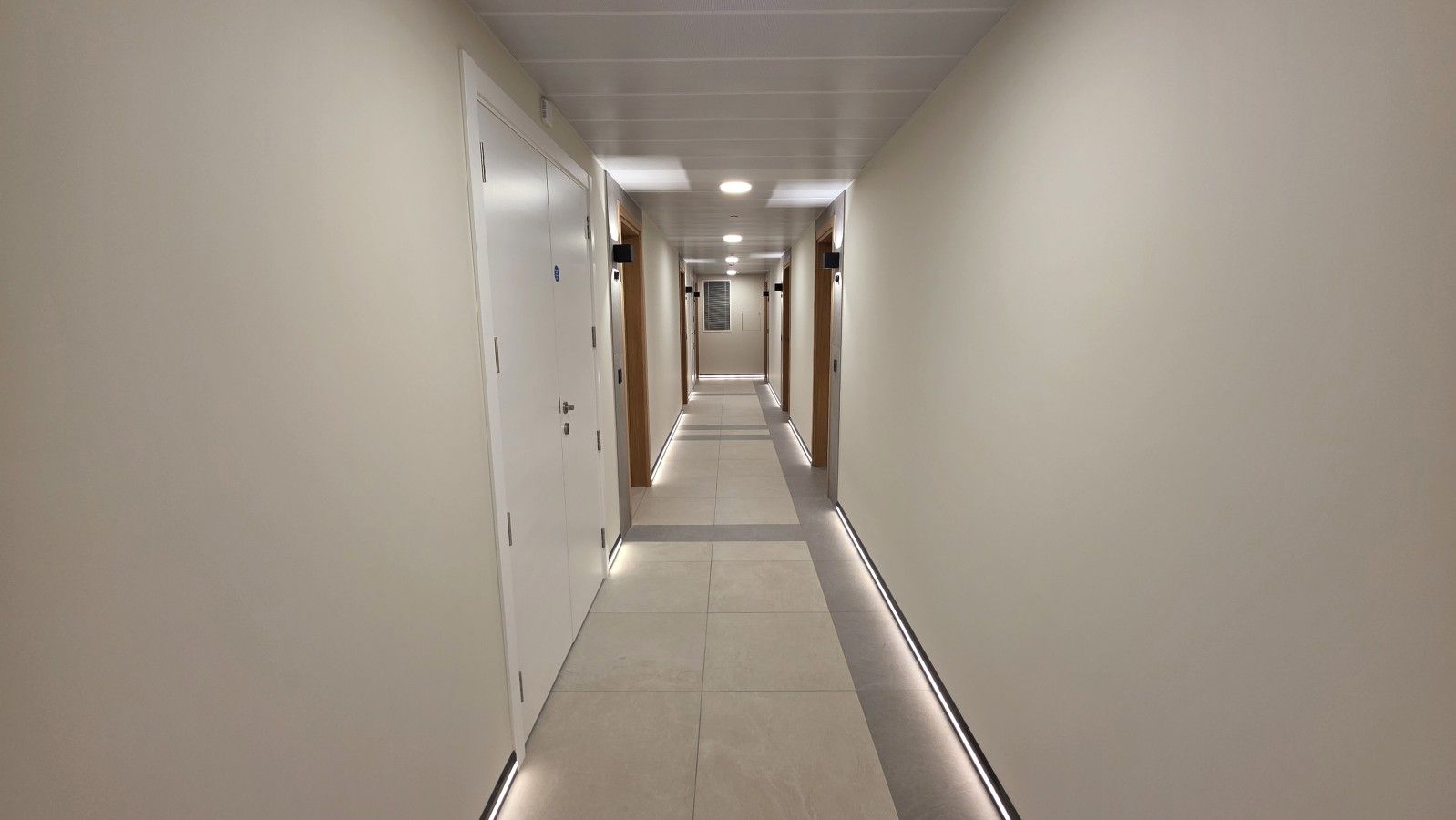 A long, narrow hotel corridor with doors on both sides and a tiled floor.