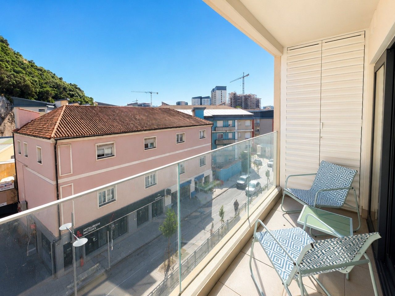 A balcony with a glass railing, two patterned chairs, and a view of buildings and a hill.