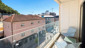 A balcony with a glass railing, two patterned chairs, and a view of buildings and a hill.
