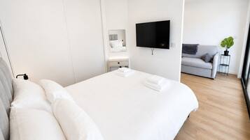 A modern hotel room with a large bed, a flat-screen TV, a grey sofa, and a small table with a plant.