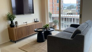 A modern living room with a flat-screen TV, a wooden console, a grey sofa, and a view of a building through large windows.