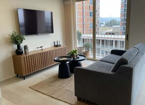 Ein modernes Wohnzimmer mit einem Flachbildfernseher, einer Holzkonsole, einem grauen Sofa und Blick auf ein Gebäude durch große Fenster.
