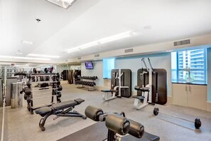 Ein Fitnessraum mit diversen Trainingsgeräten, einem Fernseher und einem Fenster mit Blick auf Gebäude.