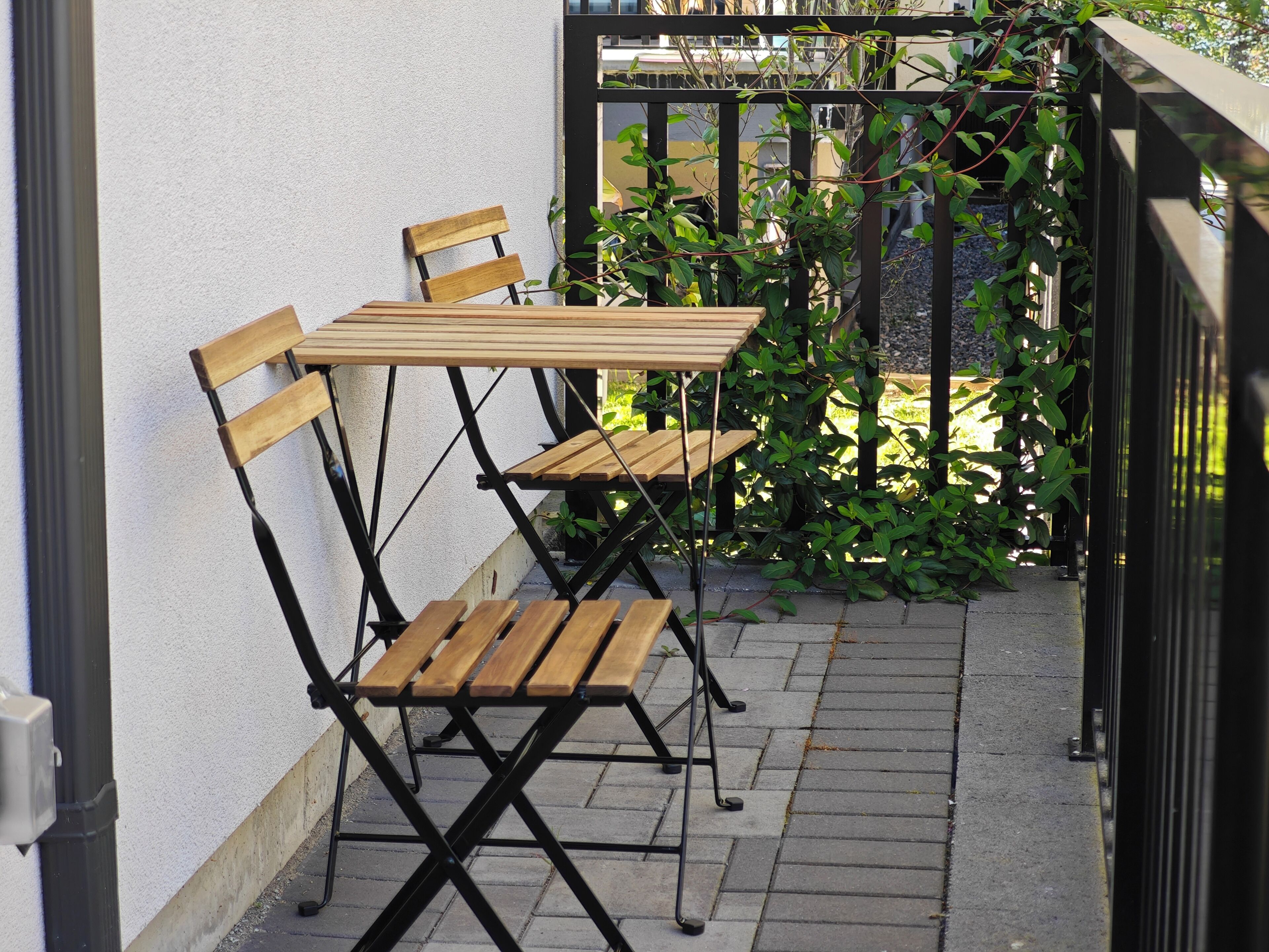 A small outdoor seating area with a wooden table and chairs, a black metal railing, and green foliage.