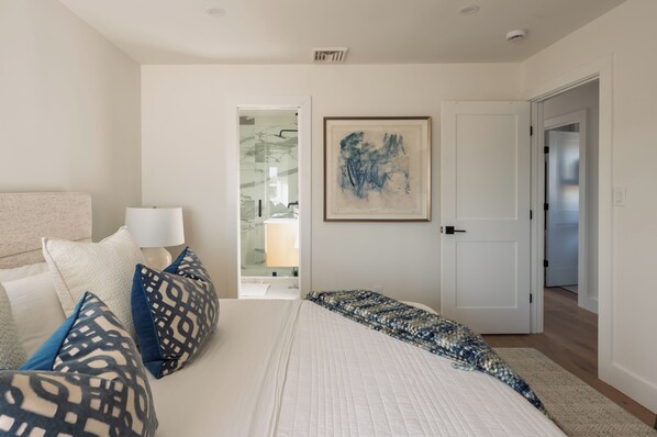 Un dormitorio con cama, almohadas, una lámpara, un cuadro abstracto enmarcado y una puerta que da a otra habitación.