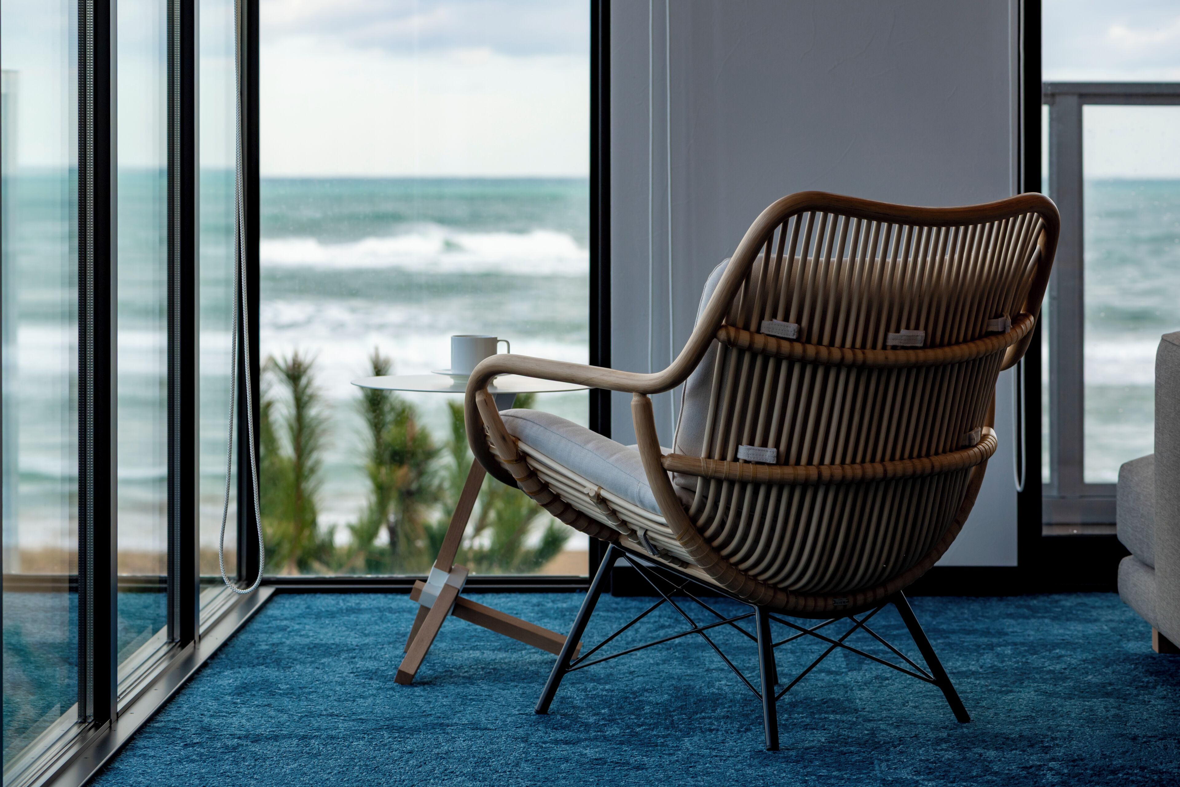 A wicker chair with a cushion, a small table with a cup, and a view of the ocean through large windows.