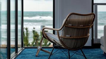 A wicker chair with a cushion, a small table with a cup, and a view of the ocean through large windows.