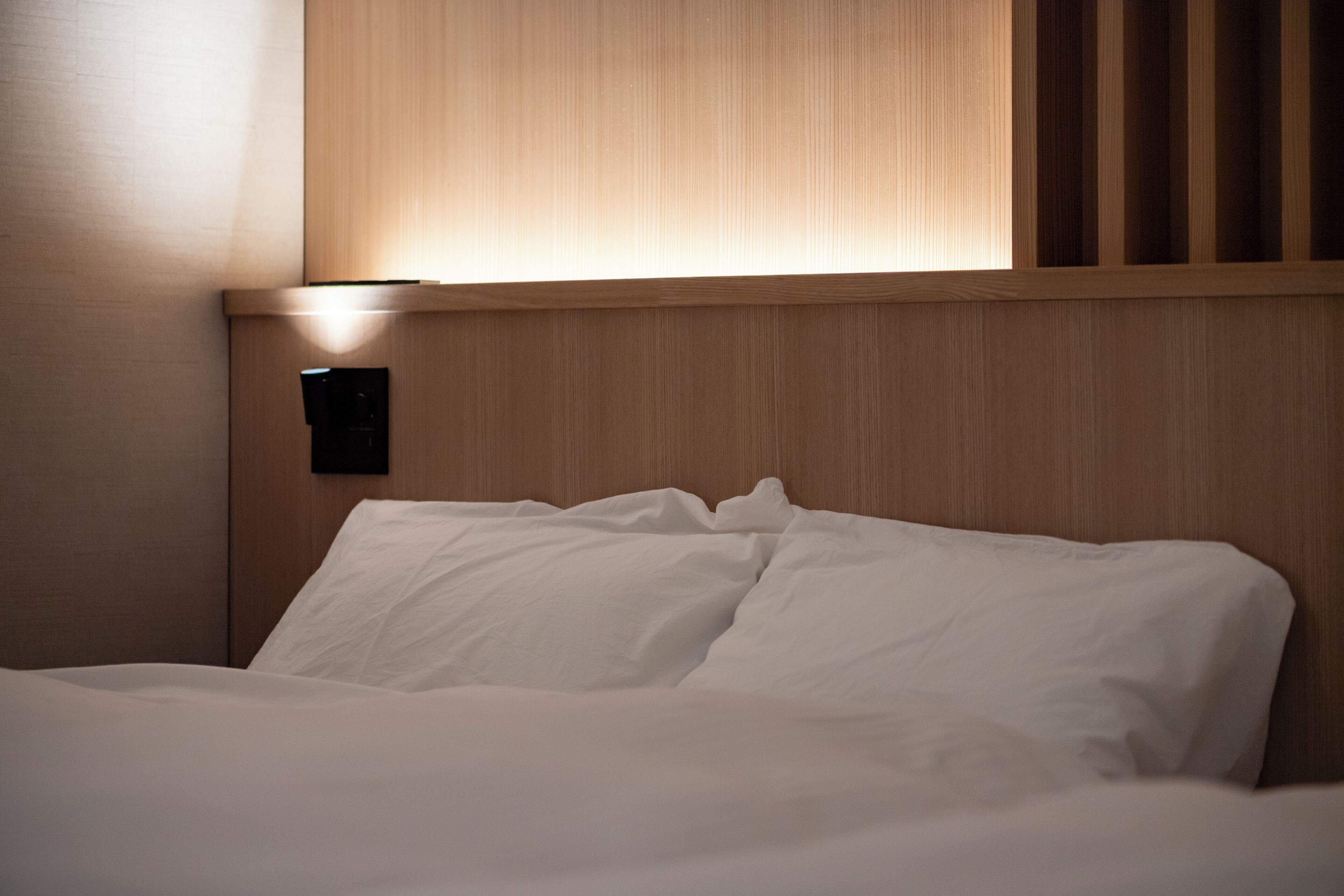 Una cama bien hecha con sábanas blancas, cabecera de madera y lámpara fijada a la pared.