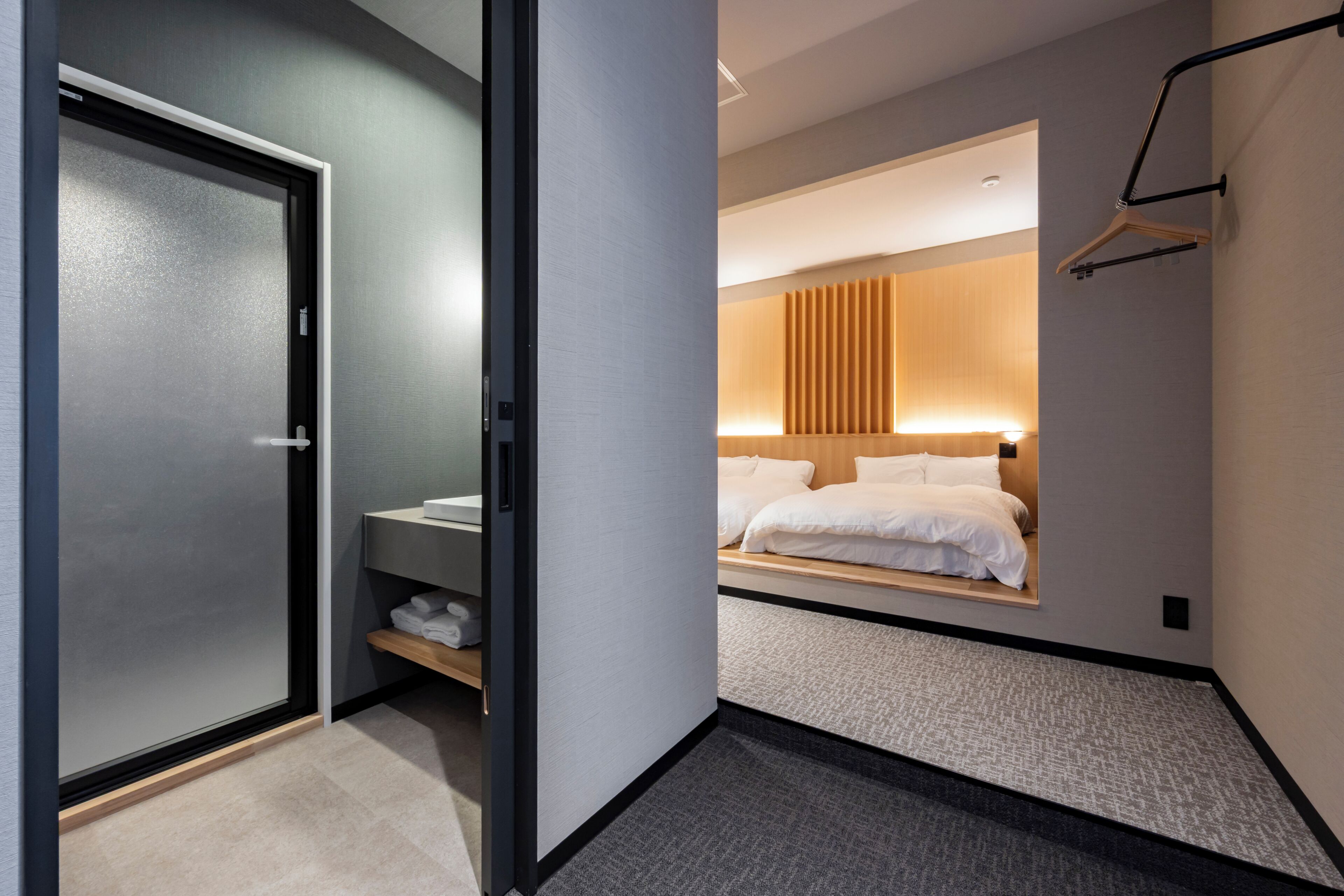A modern hotel room with a sliding door, a bedside table, a bed with white linens, a hanging light fixture, and a wall-mounted coat rack.