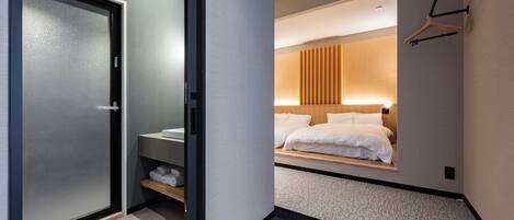 A modern hotel room with a sliding door, a bedside table, a bed with white linens, a hanging light fixture, and a wall-mounted coat rack.
