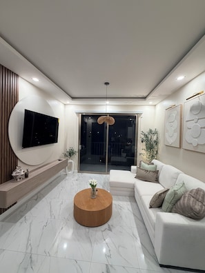 A modern living room with a flat-screen TV, a round wooden coffee table, a white sofa, and a large window.