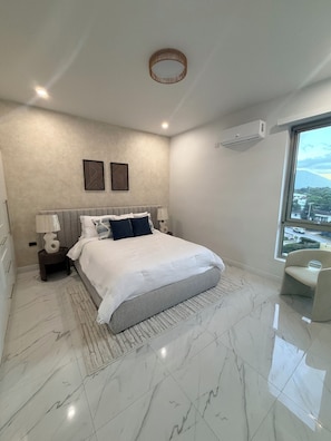 A modern bedroom with a large bed, a bedside table with a lamp, a wall-mounted air conditioner, and a large window offering a view of the outside.