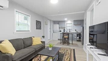 Een moderne woonkamer met een grijze bank, een salontafel en een televisie. Achtergrond toont een keuken met witte kasten en een eethoek.