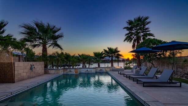 Piscina con sillas de descanso y sombrillas, rodeada de palmeras y vistas al atardecer.