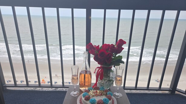 A table with a birthday cake, a vase of red roses, and two glasses of champagne on a balcony overlooking the beach. - Oceanfront Nautical Escape. Pools, Lazy River & Stunning Views (Myrtle Beach)