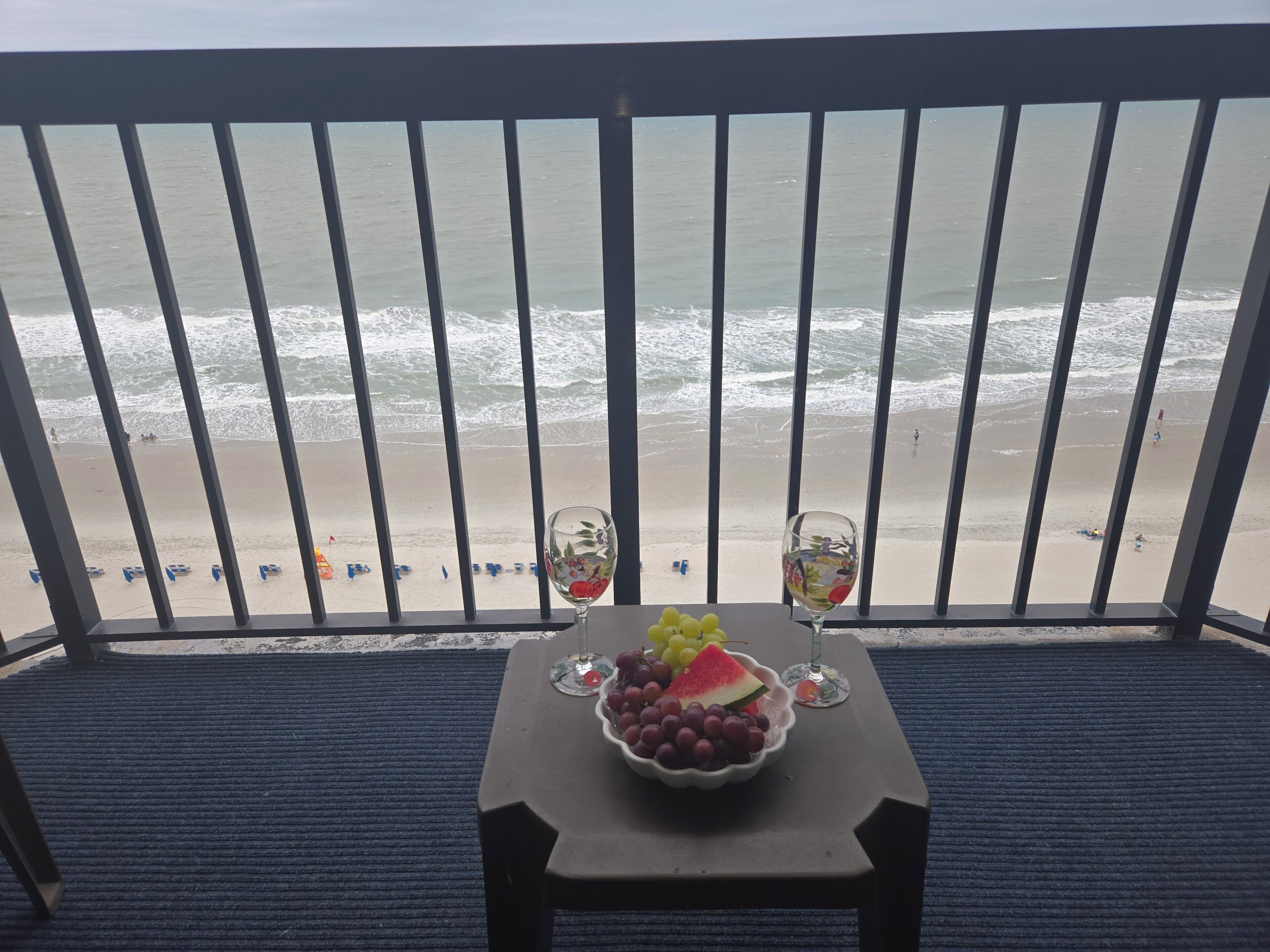 A balcony with a view of the beach, a table with a fruit bowl and two glasses.