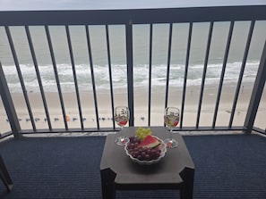 A balcony with a view of the beach, a table with a fruit bowl and two glasses. - Oceanfront Nautical Escape. Pools, Lazy River & Stunning Views (Myrtle Beach)