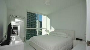 Ein Schlafzimmer mit Bett, Schreibtisch mit Computer, Fenster mit Blick auf Gebäude und einer Tür zu einem anderen Raum.