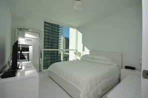 Ein Schlafzimmer mit Bett, Schreibtisch mit Computer, Fenster mit Blick auf Gebäude und einer Tür zu einem anderen Raum.